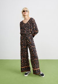 Combinaison florale noire avec de vives motifs orange et verts, à la coupe ample, avec des jambes larges et une taille cintrée. Associée à des baskets.