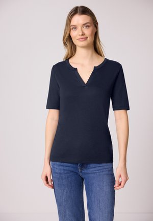 Jeune femme portant un haut bleu marine à manches courtes avec un décolleté en V et un jean bleu, debout devant un fond clair uni.