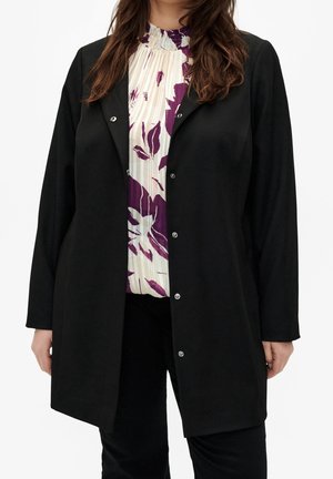 Femme portant un manteau noir boutonné sur un chemisier blanc à imprimé floral violet, associé à un pantalon noir, debout devant un fond blanc.