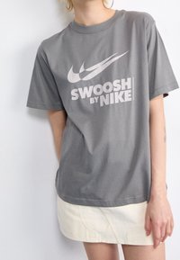 Šedé bavlněné tričko s krátkým rukávem, které na přední straně obsahuje velké bílé logo "Swoosh" a text "SWOOSH BY NIKE" tučným písmem.