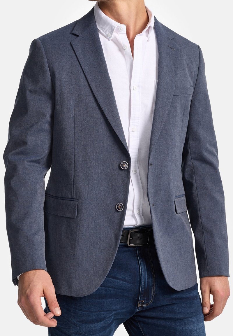 Dunkelblauer Blazer aus strukturiertem Stoff, mit zwei Knöpfen, Kerbkragen und einer Brusttasche, getragen über einem weißen Hemd und Jeans.