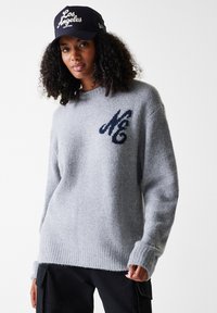 Grå strikket genser med ribbestrikket kant og mansjetter, med en mørk blå "MC"-logo. Kombinert med en svart caps med hvit "Los Angeles"-tekst.