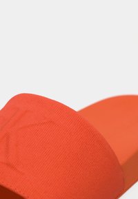 Nærbillede af en orange tekstureret stof slidesandal med en bred rem over foden og en flad sål.