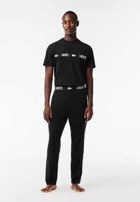 Schwarz Baumwoll-T-Shirt mit weißen Lacoste-Logos, kombiniert mit schwarzen Hosen, die einen weißen Lacoste-Hosensaum haben. Einfaches, figurbetontes Design.
