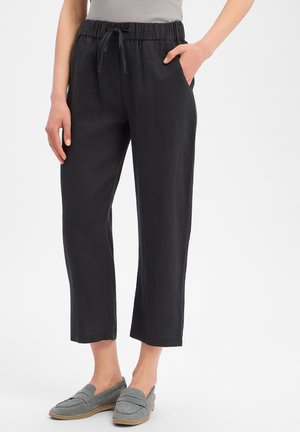 Vrouw draagt zwarte korte broek met trekkoord en zijzakken en grijze instapschoenen, staande tegen een effen achtergrond.