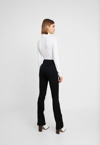 Kvinne med kort brunt hår som har på seg hvit høyhalset genser, svarte høytaljede buksesvetter og hvite ankelboots med hæl, stående mot en ensfarget bakgrunn.