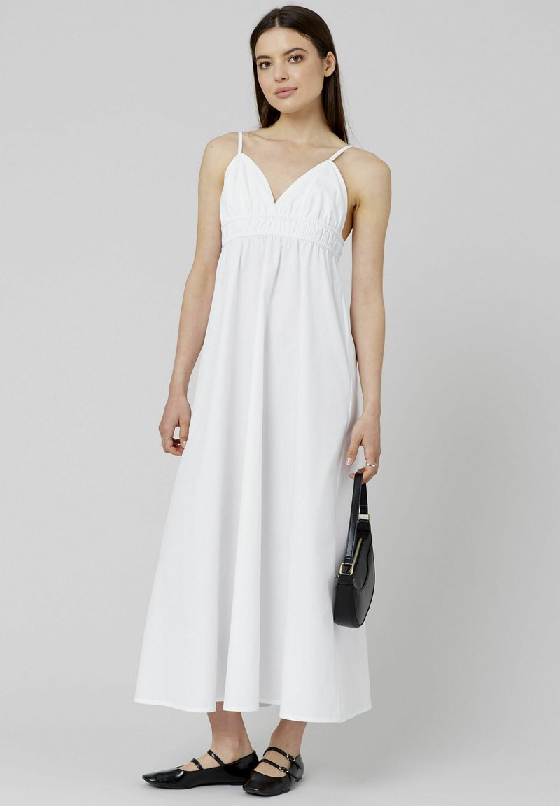 Robe maxi blanche avec des fines bretelles, taille froncée et jupe fluide. Associée à des chaussures plates noires et un petit sac à main noir.