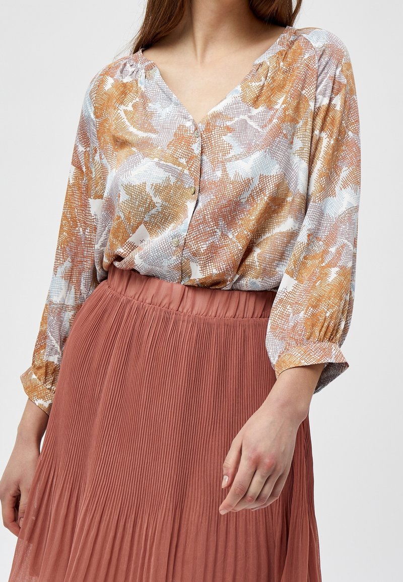 Blusa in tessuto fantasia con tonalità beige, arancione e blu, con frontale a bottoni. Abbinata a una gonna plissettata rosa pallido.