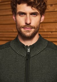 Tmavě zelená zipová bunda s texturovaným pleteným vzorem, žebrovaným límcem a kontrastní hladkou tkaninou na ramenou. Viditelné detaily zipu.