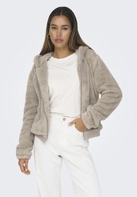 Giacca zip-up beige chiaro foderata in pelliccia con cappuccio, caratterizzata da polsini e orlo a coste. Indossata sopra una camicia bianca e abbinata a pantaloni bianchi.