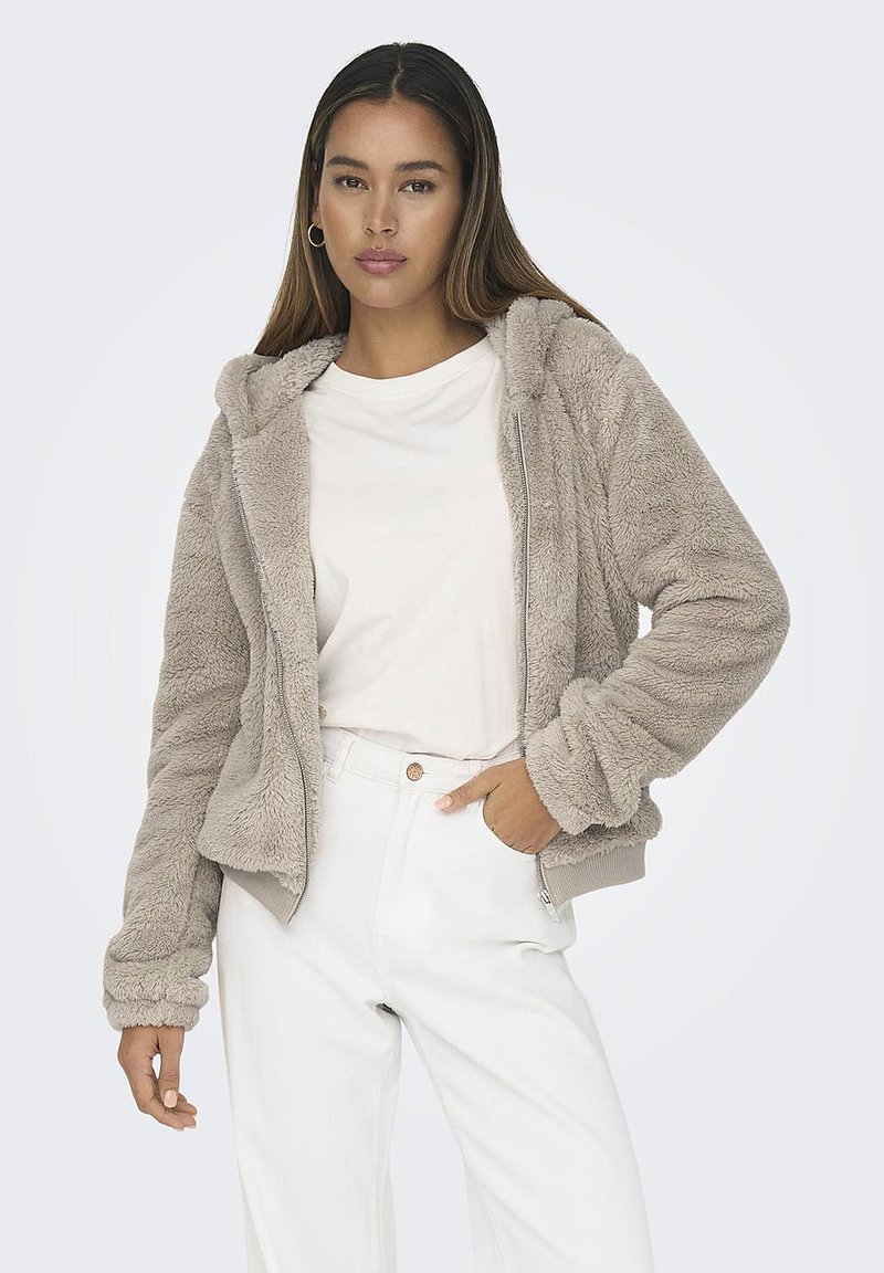 Giacca zip-up beige chiaro foderata in pelliccia con cappuccio, caratterizzata da polsini e orlo a coste. Indossata sopra una camicia bianca e abbinata a pantaloni bianchi.