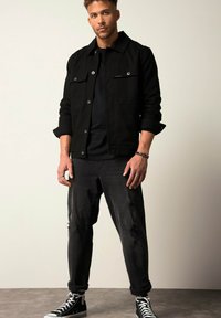 Zwarte denimjas met een knoopsluiting en dubbele borstzakken, gecombineerd met versleten zwarte jeans en zwarte high-top sneakers.