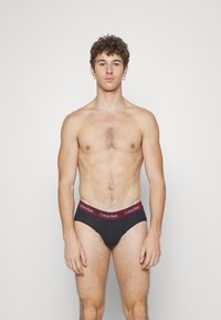 Schwarze Baumwoll-Bikini-Briefs mit einem bordeauxfarbenen Elastikbund, der das "Calvin Klein"-Logo trägt. Glatte Textur, enganliegendes Design und niedriger Schnitt.