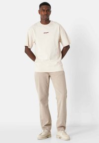 Kremowy bawełniany t-shirt z logo na piersi, zestawiony z jasnobeżowymi spodniami i beżowymi butami. Prosty design i luźny krój.