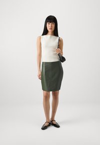 Zöld bőr ceruzaszoknya egy szűk, fehér bordázott toppal párosítva. A modell egy kis fekete táskát tart, fekete lapos cipőt visel. Egyszerű, modern dizájn.