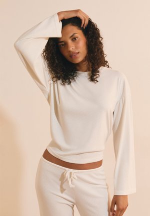 Donna con capelli ricci che indossa una maglia bianca a maniche lunghe e pantaloni bianchi coordinati, posando con una mano sulla testa su uno sfondo beige.