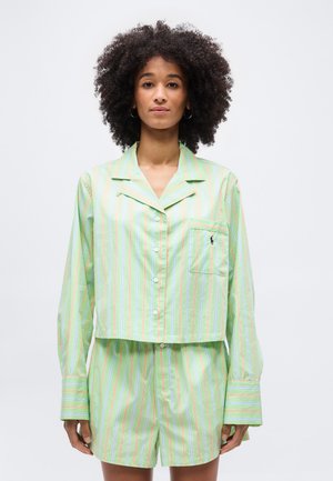 Woman with curly hair wearing light green, striped long-sleeve button-up shirt and matching shorts against a plain white background.