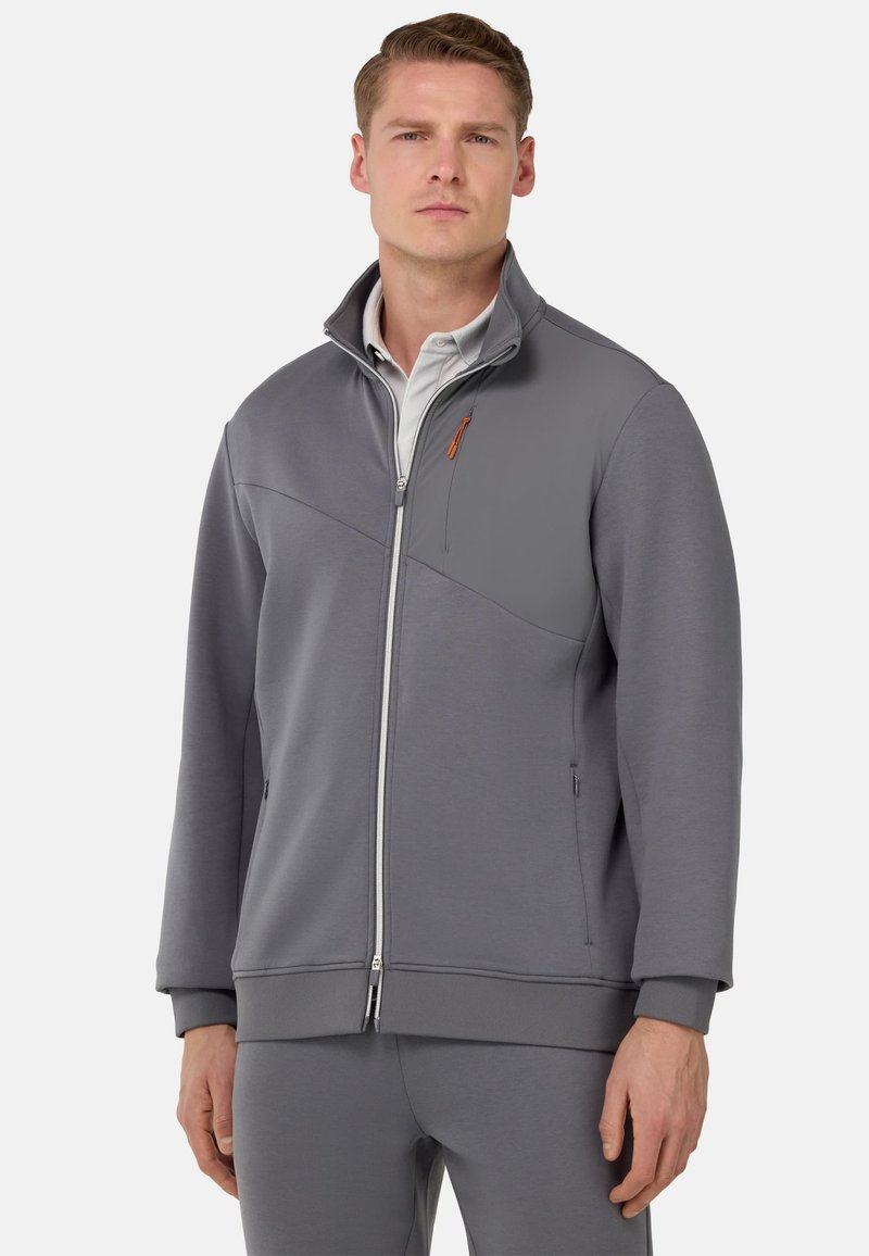 Veste zippée grise avec col montant et panneau contrastant. Comprend des coutures visibles, des poches latérales et un accent orange subtil sur la fermeture éclair.