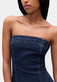 Tamnoplava traper haljina bez naramenica s uskim krojem, vidljivim šavovima i ravnim izrezom. Glatka tekstura s blagim sjajem.