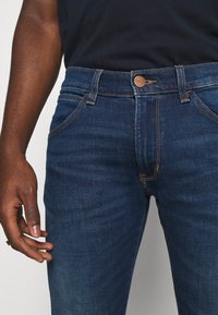 Jeans in denim blu scuro con un design aderente, caratterizzati da cuciture a contrasto, un piccolo logo sul bottone e una vestibilità a cinque tasche.