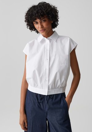 Jeune femme aux cheveux bouclés portant une chemise blanche sans manches à boutons et un pantalon bleu foncé à cordon, debout avec une main dans la poche.