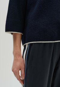 Marineblaues Rippenstrickoberteil mit dreiviertel Ärmeln und weißem Saumakzent. Getragen mit schwarzen Hosen, die seitliche Streifen in Hellblau und Weiß haben.