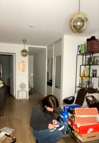 Persoană așezată pe podea leagă șireturile, înconjurată de cutii de încălțăminte stivuite și rafturi cu cărți și decorațiuni într-un living.