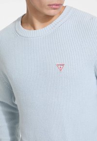 Pull en tricot bleu clair avec un col ample, texture côtelée et petit logo triangulaire rouge sur la poitrine gauche. Matière en mélange de coton.