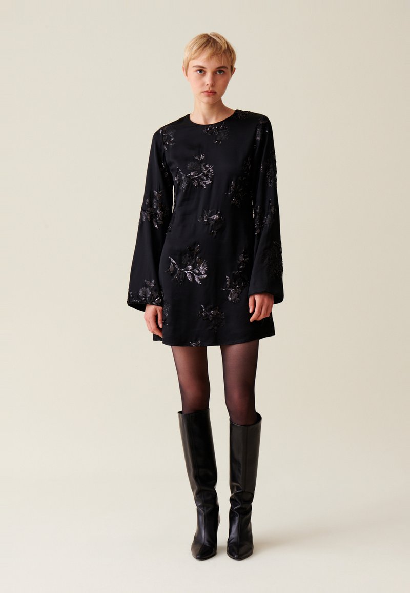 Schwarzes Minikleid mit langen Ärmeln, verziert mit floralen Mustern. Aus glattem Stoff gefertigt, kombiniert mit kniehohen schwarzen Stiefeln.