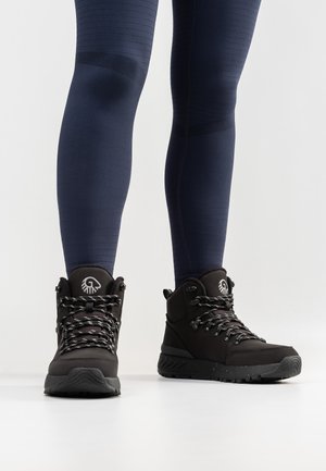 Schwarze Wanderschuhe mit strukturiertem Äußeren, grauen Schnürsenkeln und einem markanten Logo. Getragen mit dunkelblauen, figurbetonten Leggings.
