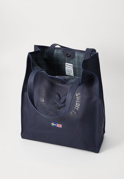 Marineblå totebag laget av glatt materiale, med et sirkulært emblem og svenske og britiske flagg på fronten. Fôret med et rutete mønster.