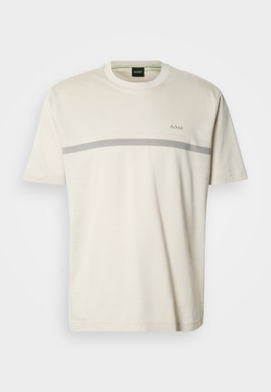 T-shirt beige clair en coton doux, avec une bande horizontale grise traversant la poitrine, et un logo brodé au-dessus de la bande.