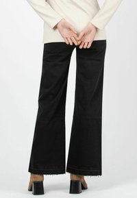 Schwarze Schlagjeans mit fransigem Saum, aus Denimstoff; kombiniert mit einem beigen Oberteil und braunen Absatzschuhen, von hinten betrachtet.