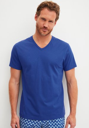 Blaues V-Ausschnitt-T-Shirt aus Baumwolle, kurzärmelig, mit glatter Textur. Getragen mit gemusterten Badeshorts mit Wellenmustern.