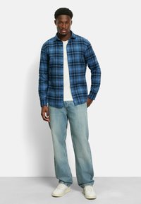 Blau kariertes Flanellhemd mit knöpfbarem Vorderteil, getragen über einem weißen T-Shirt. Lässige hellblaue Jeans und weiße Sneaker runden das Outfit ab.