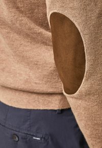 Pull en maille beige avec patch en suède brun au niveau du coude, porté avec un pantalon bleu foncé, montrant des coutures détaillées et une texture apparente.