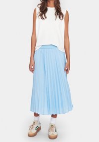 Harmaankappaleet sininen laskostettu hame, jossa on sileä tekstuuri, yhdistettynä hihattomaan valkoiseen toppiin ja vaaleanhirveisiin tennareihin, joissa on vihreät raidat.