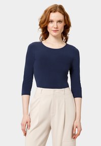 Tmavě modrý body s dlouhým rukávem, kulatým výstřihem, hladkou texturou a přiléhavým střihem, doplněný o světle béžové kalhoty s vysokým pasem.
