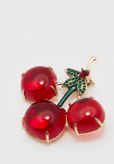 Καρφίτσα σε χρυσούς τόνους με τρία μεγάλα κόκκινα κεράσια από πολύτιμους λίθους και φύλλα από πράσινους στρας σε λευκό φόντο.