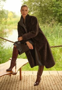 Barna plüss kabát magas gallérral, sötét bőr ruhára viselve, térdig érő barna magassarkú csizmával, természetes háttér előtt.