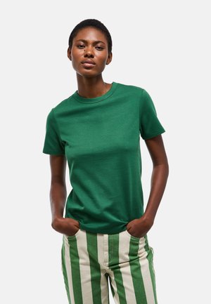 Femme portant un t-shirt vert uni à manches courtes et un pantalon rayé verticalement vert et blanc, debout, les mains dans les poches.