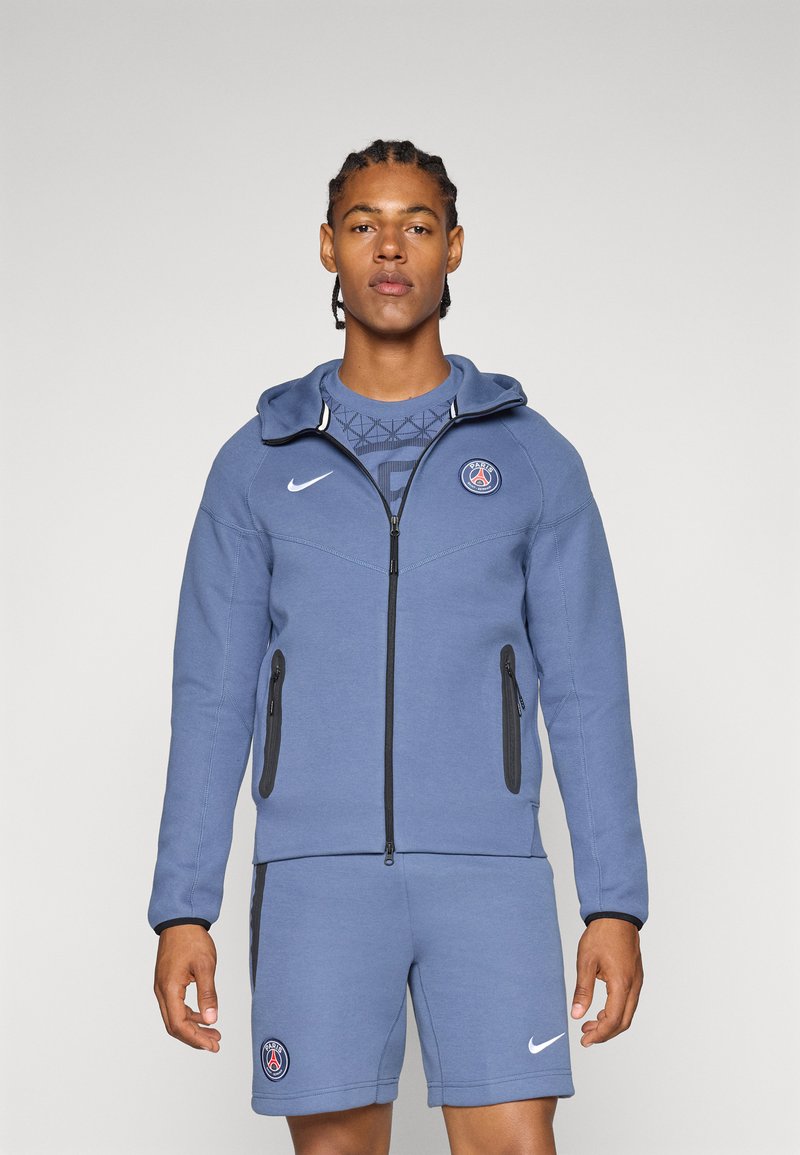 Felpa blu con zip frontale, accenti neri e tasche laterali. Indossata sopra una camicia blu coordinata a fantasia e pantaloni corti, con loghi.
