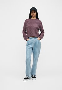 Γ υναίκα που φοράει μωβ πουλόβερ με πλέξη κοτσίδα, ανοιχτό μπλε τζιν με ρίγες, μαύρο καπέλο και μαύρα-λευκά αθλητικά παπούτσια, στέκεται με τα χέρια στις τσέπες.