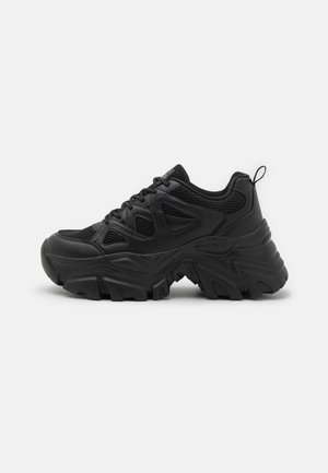 Black athletic shoes with a textured mesh upper, curved rubber sole, and prominent tread pattern. Features integrated lacing and pull tab.