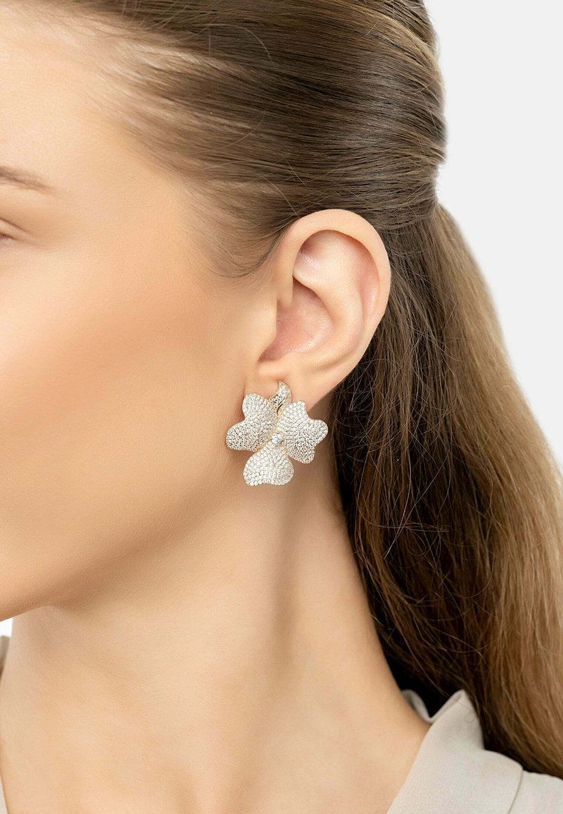 Woman’s ear with hair pulled back, wearing a large flower-shaped earring covered in small sparkling stones.