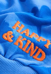 Blauer Stoff mit einer weichen Textur zeigt orangefarbene Stickereien mit dem Text "HAPPY & KIND", was ein verspieltes Designelement hervorhebt.