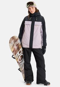 Mujer vestida con una chaqueta de nieve negra y lavanda, pantalones de nieve negros, guantes y gafas, sosteniendo una tabla de snowboard con un patrón marrón y blanco.