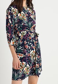 Vestido floral en azul marino oscuro con mangas cortas, cintura ceñida y un patrón de hojas y flores coloridas. Tejido suave con un ligero brillo.