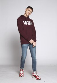 Bordová mikina s bielym logom Vans, v kombinácii s prispôsobenými modrými džínami a červenými a bielymi teniskami s vzorom. Model stojí v uvoľnenej póze.