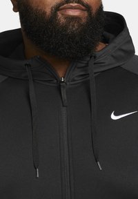 Sudadera negra de Nike con cierre de cremallera, cordones ajustables y logo blanco. Tela suave y texturizada con paneles de contraste en los hombros.